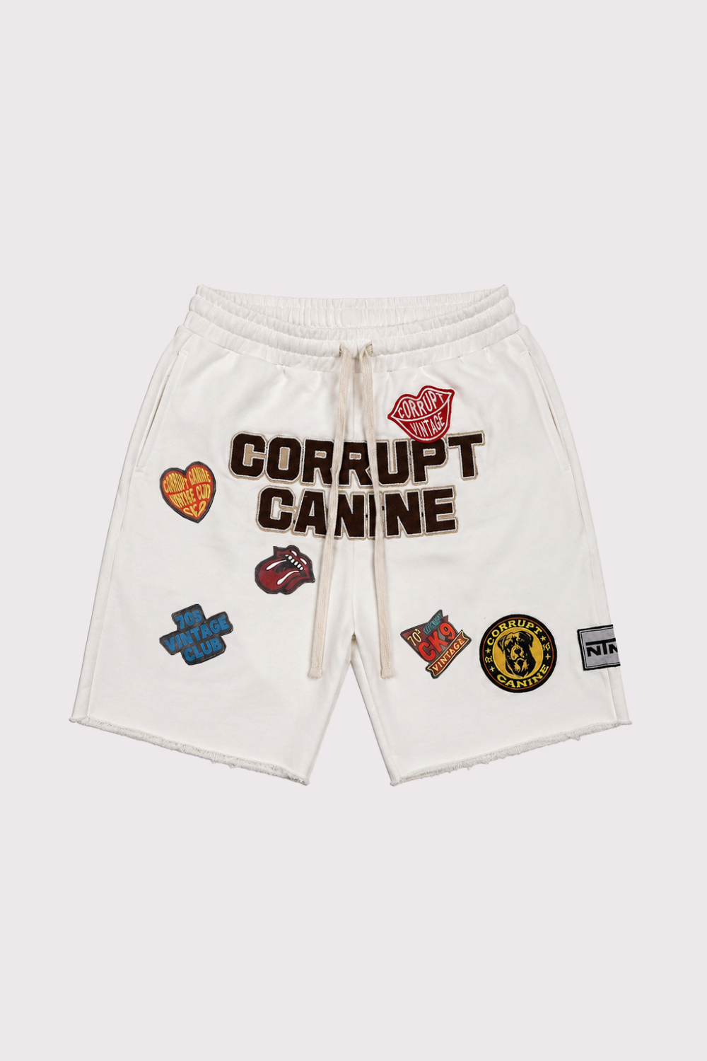 White sweat shorts with 'CORRUPT CANINE' text and multiple colorful embroidered patches, shown flat on a light gray background.