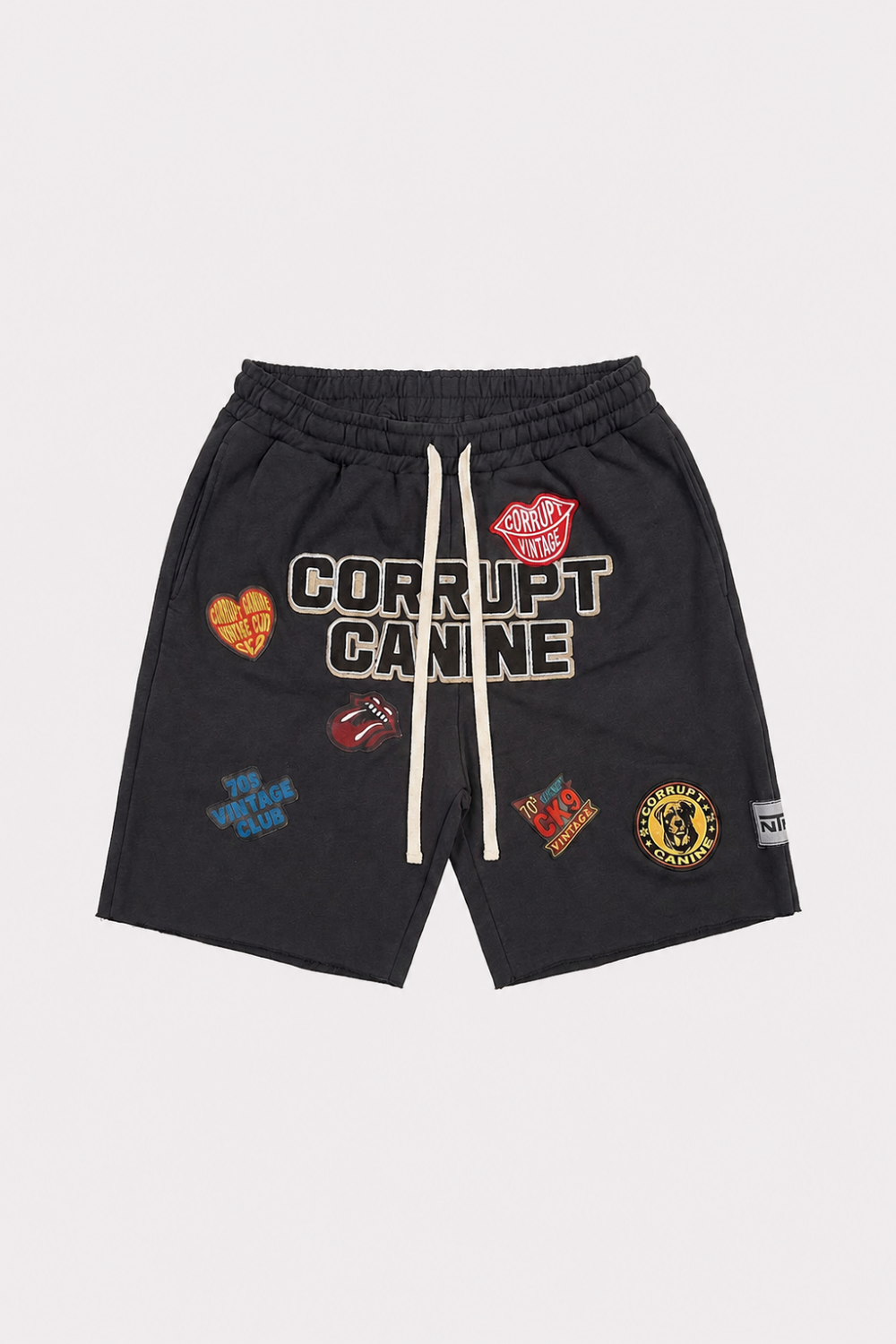Black shorts with 'CORRUPT CANINE' embroidered across the front, decorated with multiple colorful patches, and featuring cream-colored drawstrings, shown flat on a light gray background.