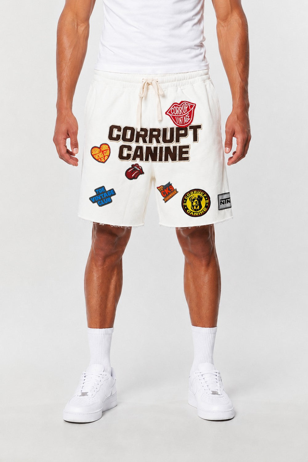 A model wearing white sweat shorts with 'CORRUPT CANINE' text and several colorful patch graphics, paired with a white t-shirt, white socks, and white sneakers against a light gray studio background.