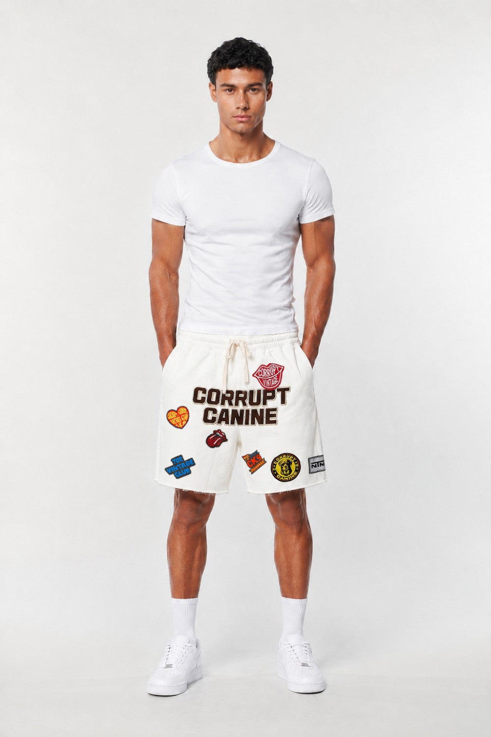 A male model wearing a white crew-neck t-shirt and white shorts with 'CORRUPT CANINE' text and colorful patches, standing against a light gray studio background.
