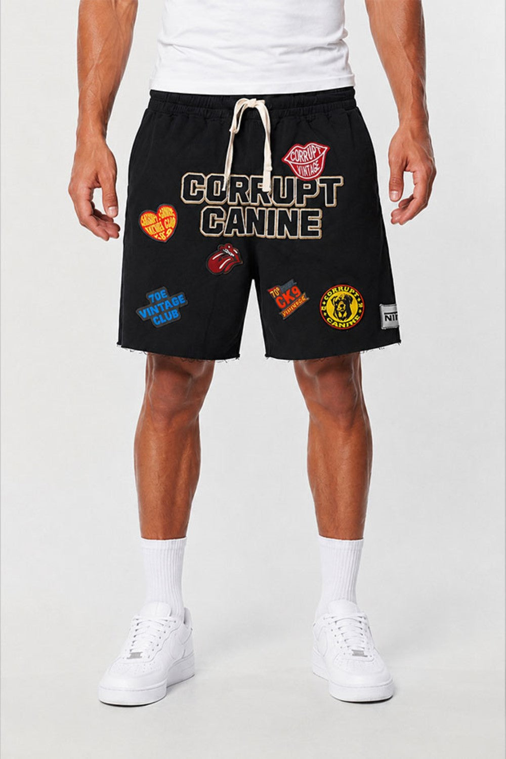 A model wearing black Corrupt Canine athletic shorts with multiple colorful patches and a white drawstring, paired with a white t-shirt, white socks, and white sneakers against a light gray studio background.
