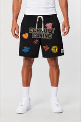 A model wearing black Corrupt Canine athletic shorts with multiple colorful patches and a white drawstring, paired with a white t-shirt, white socks, and white sneakers against a light gray studio background.