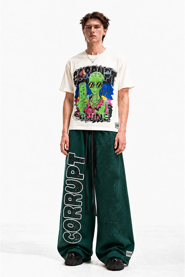 Man wearing a cream graphic t-shirt with an alien design and green wide-leg pants with "CORRUPT" printed vertically.