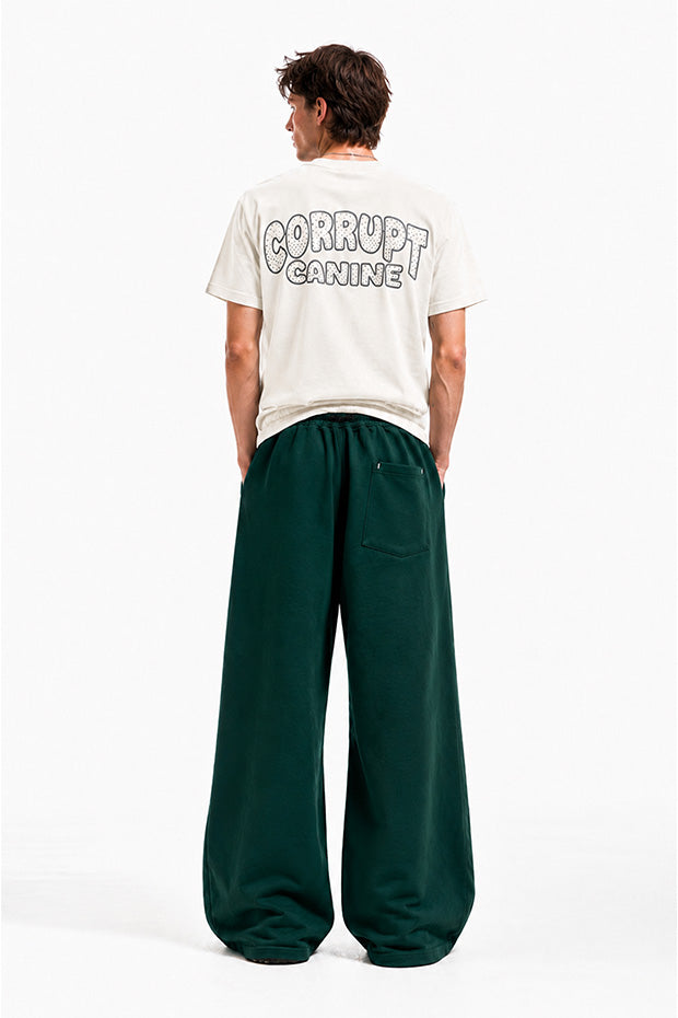 Man wearing a cream graphic t-shirt with "CORRUPT CANINE" text and dark green wide-leg sweatpants.