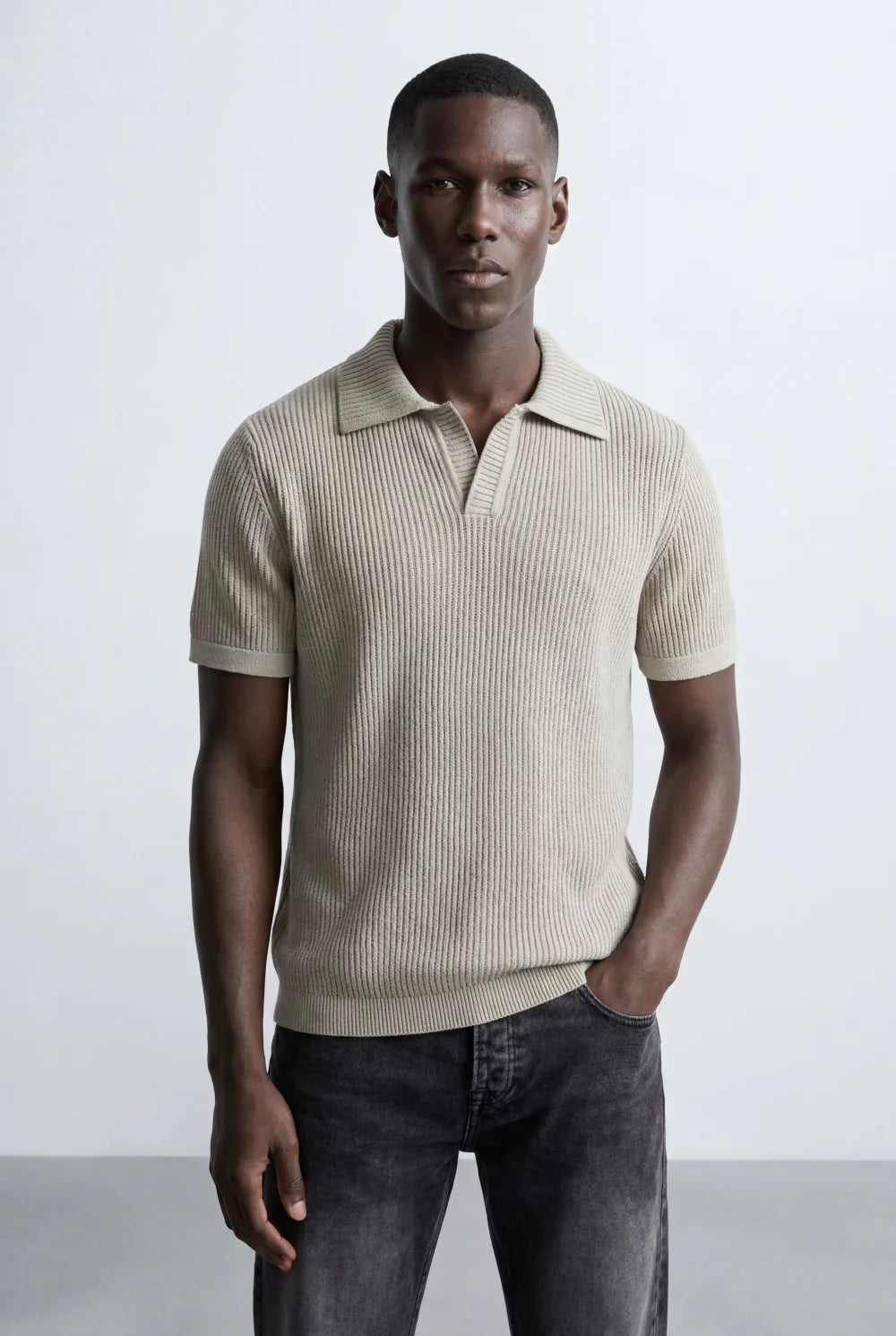 Man wearing a beige ribbed knit short-sleeve polo shirt with an open collar and dark wash jeans.
