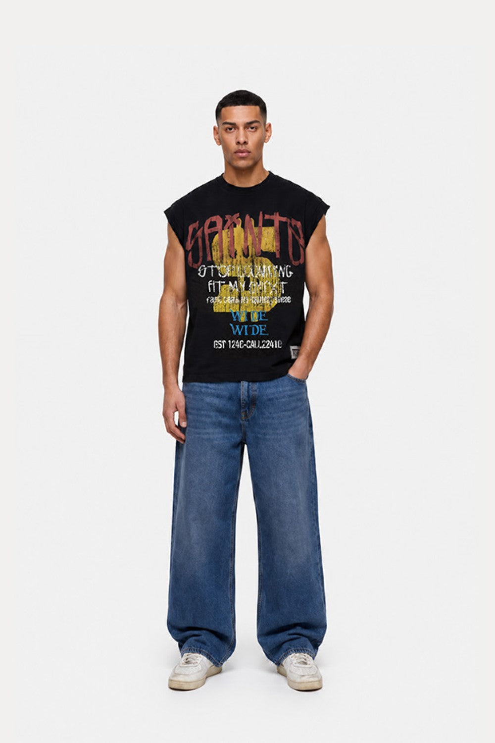A male model stands against a light gray studio background wearing a black sleeveless graphic T-shirt with colorful text and blue wide-leg jeans.