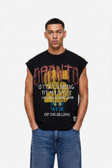 A man wearing a sleeveless black t-shirt with a colorful graphic print and blue jeans, standing against a plain white background.