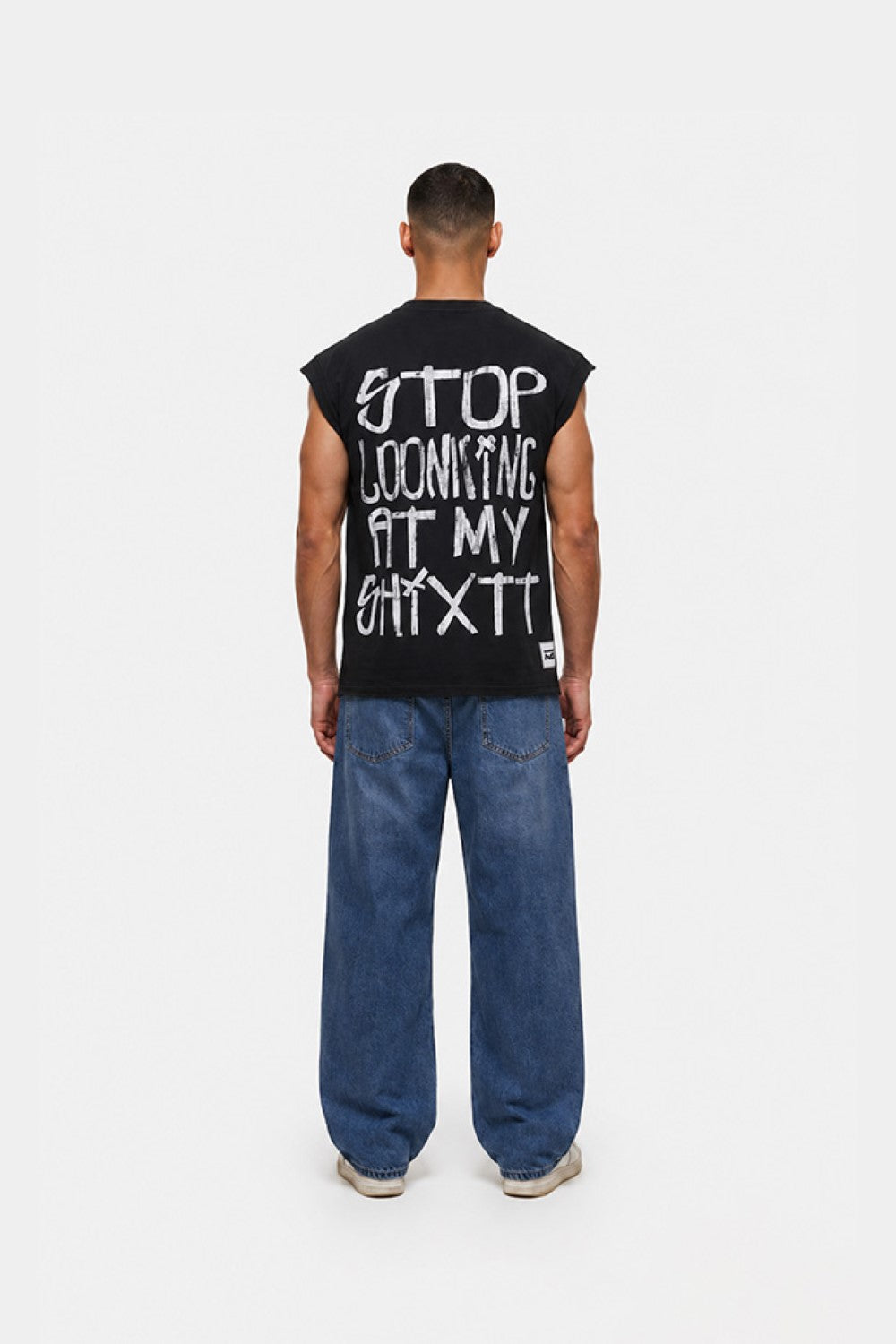 A man viewed from behind wearing a black sleeveless t-shirt with white graphic text on the back, paired with loose blue jeans and white sneakers, against a plain white studio background.