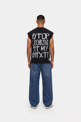A man viewed from behind wearing a black sleeveless t-shirt with white graphic text on the back, paired with loose blue jeans and white sneakers, against a plain white studio background.